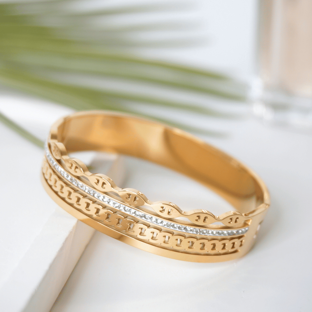 Gold bracelet with intricate design on a white surface with green leaves in the background